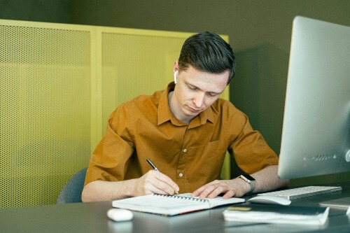 Man sitting in front of computer, taking notes in a notebook